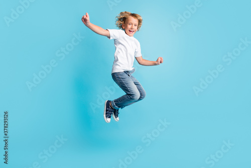 Small kid jump raise hands excited smile wear white shirt jeans sneakers isolated blue color background