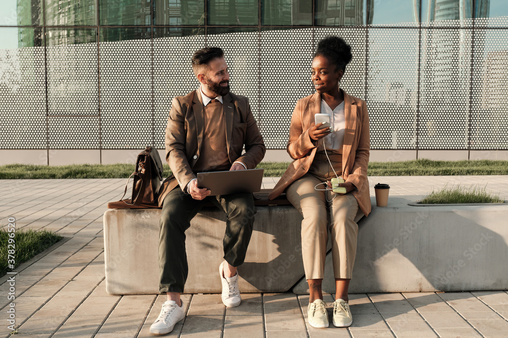 Two business colleagues sitting on bench and talking to each other outdoors