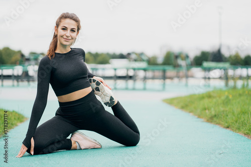 portrait of a happy woman in black sports clothes looking at the camera and smiling. does leg stretching before sports training doing sports