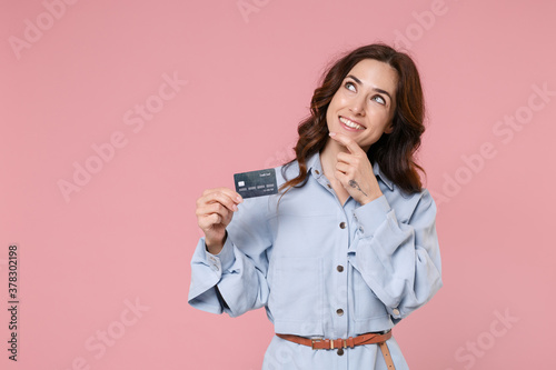 Pensive young brunette woman 20s wearing casual blue shirt dress posing holding in hand credit bank card put hand prop up on chin looking up isolated on pastel pink colour background, studio portrait.