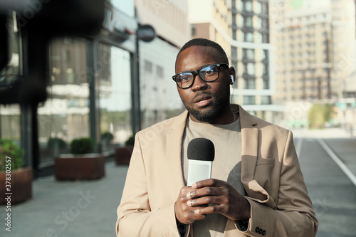 Portrait of African journalist in eyeglasses speaking in microphone while working in the city