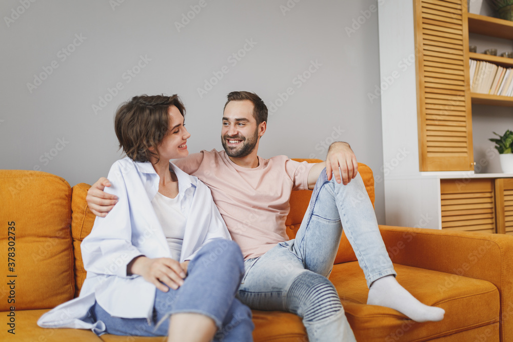 Smiling cheerful pretty beautiful young couple two friends man woman 20s wearing casual clothes sitting on couch hugging looking at each other resting relaxing spending time in living room at home.