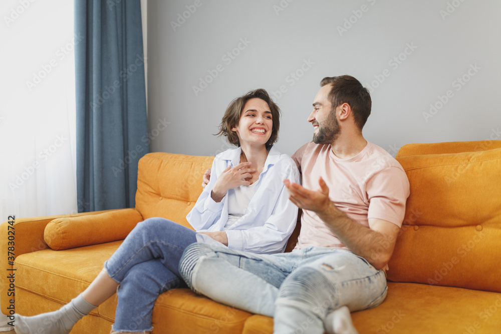 Smiling laughing funny young couple two friends man woman 20s in casual clothes sitting on couch hugging speaking talking looking at each other resting relaxing spending time in living room at home.
