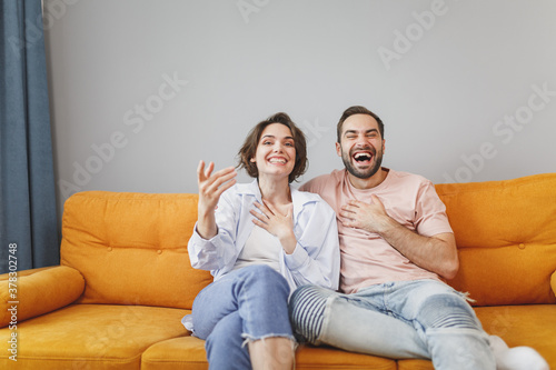 Laughing cheerful funny young couple two friends man woman 20s wearing casual clothes sitting on couch hugging spreading hands looking camera resting relaxing spending time in living room at home.