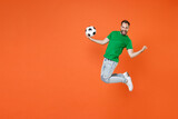 Full length portrait happy man football fan in green t-shirt cheer up support favorite team with soccer ball jumping doing winner gesture isolated on orange background. People sport leisure concept.