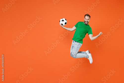 Fototapeta Naklejka Na Ścianę i Meble -  Full length portrait happy man football fan in green t-shirt cheer up support favorite team with soccer ball jumping doing winner gesture isolated on orange background. People sport leisure concept.