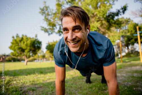 Smiling handsome young male athlete planking exercise in sunny outdoor park ear phones playing music