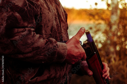 hunter with a gun refills cartridges against the background of the lake and the bright sunset in autumn © Мария Шевцова