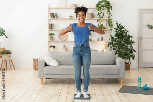 Cheerful African Girl Gesturing Thumbs-Up Standing On Weight Scales Indoor © Prostock-studio