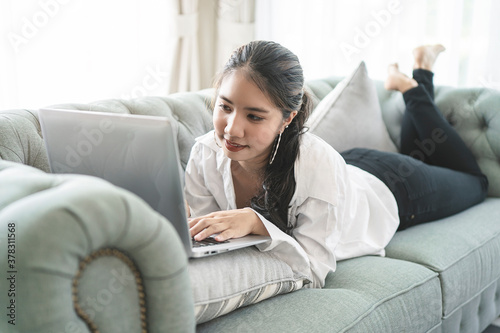 Happy casual beautiful Asian woman working on a laptop sitting on the couch in the house.