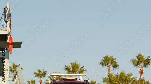 Iconic retro wooden lifeguard watch tower, baywatch red car. Life buoy, american state flag and palm trees against blue sky. Summertime california aesthetic, Santa Monica beach, Los Angeles, CA USA.