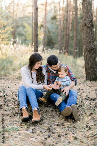 Picture of lovely parents with little kid son, in autumn park or forest, sitting together and enjoying their family time, playing outdoors. Happy family in autumn nature