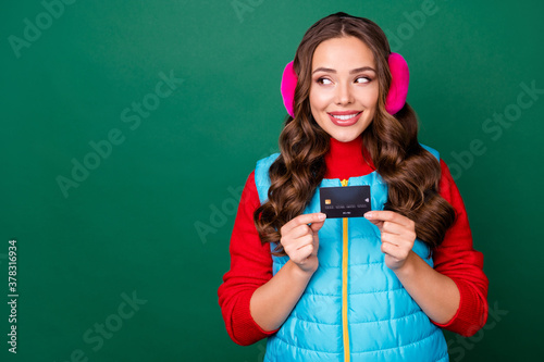 Photo of cute charming young lady hold credit card interested look copyspace presenting good variant smiling wear pink ear warmers blue vest red sweater isolated green color background