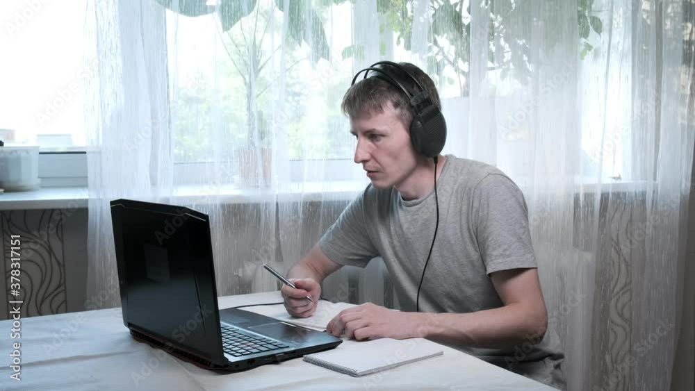 Serious male student wears headphones, studies with internet teacher at home. focused young man talks to tutor via laptop, writes notes. online education, video call tutoring, distant work, remote job