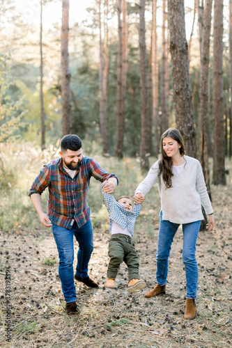 Happy joyful family, handsome bearded father, pretty brunette mother and adorable cute baby son, holding hands and walking in autumn pine forest with pine trees on the background