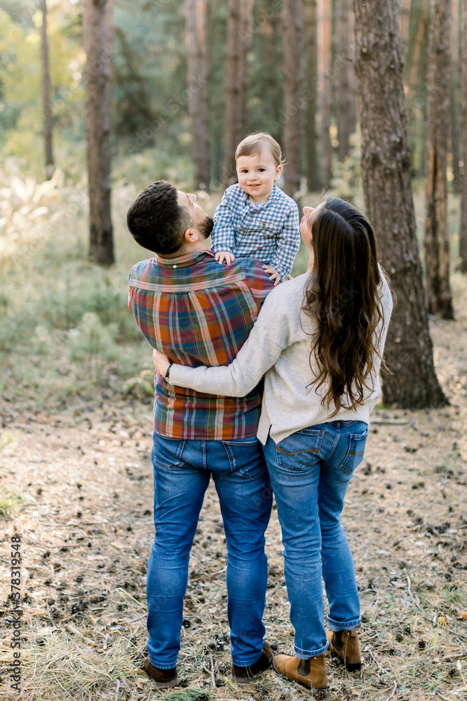 Fototapeta premium Back view of young stylish parents, father and mother, holding their little baby child boy, smiling at camera. Outdoor family portrait. Family day concept, walk with child
