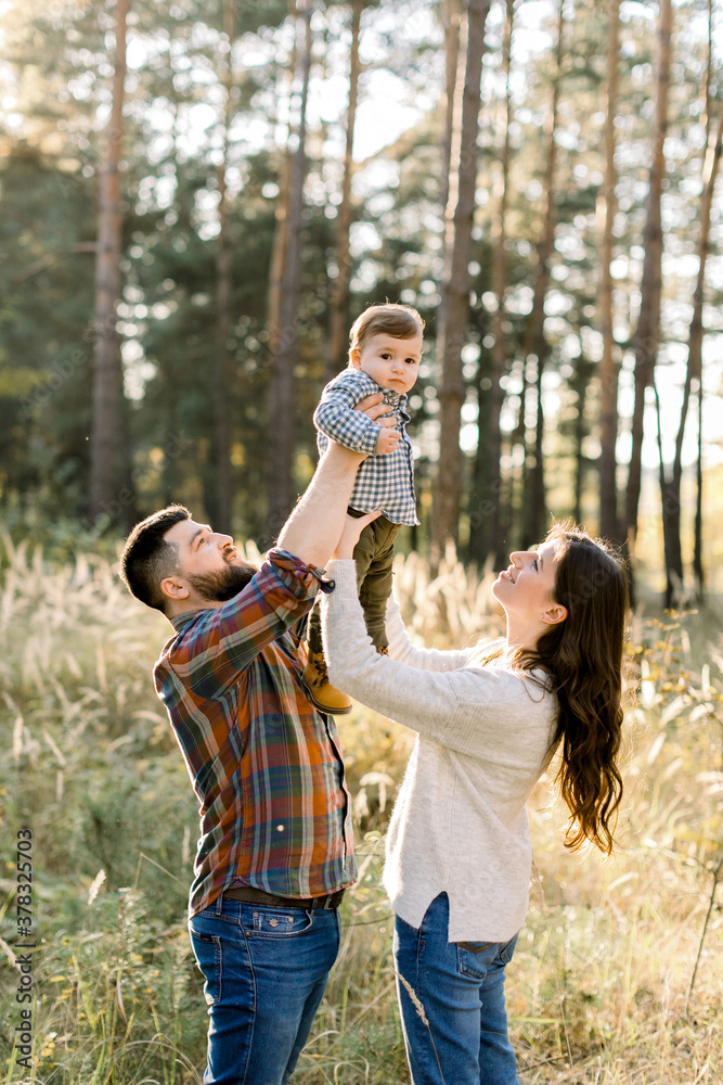 Outdoor family portrait of happy young parents, wearing stylish casual clothes, having fun and lifting up their little cute baby son, during walk in autumn forest at sunny day