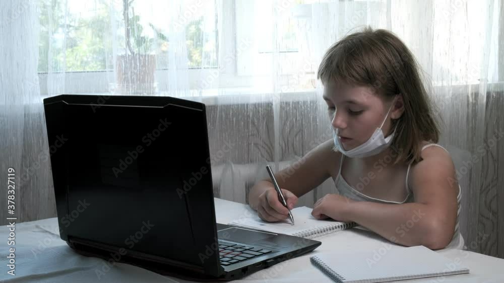 little girl wears protective mask, studies at home with internet teacher, writes notes in notebook, doing homework. schoolgirl listens to tutor, looks at laptop. online education, video call tutoring