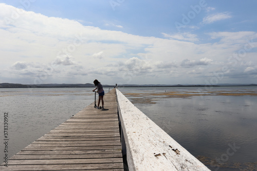 Silhouette of young girl with dark hair alone enjoying rides on a scooter in this wooden jetty bridge. Skyline background in this quite lake landscape in the Central Coast Australia © Irene's Stock