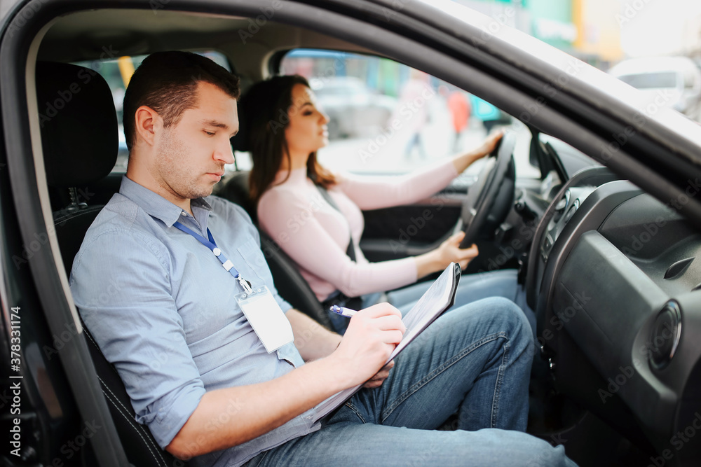 Male auto instructor takes exam in young woman. Busy, serious and concentrated man writing test results on paper. Confident female driver look forward on road. Male auto instructor takes exam in young woman. Busy, serious and concentrated man writing test results on paper. Confident female driver look forward on road.