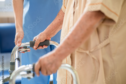 Close up of elderly patient hands using walking frame with nurse assistance. Medical professional or caregiver assisting senior man to walk with walker during recovery in hospital. 