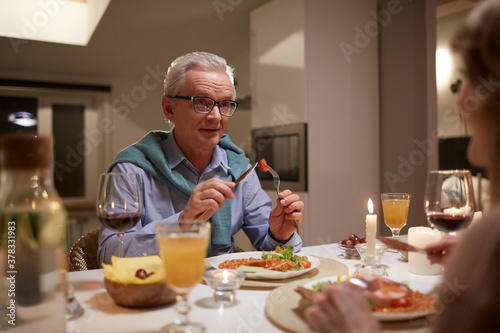Senior man sitting at the table eating meal and talking to his wife during their family dinner at home