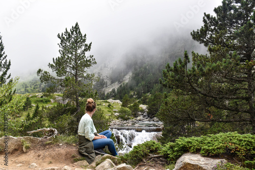 Woman sitting on a rock looking at a river of turquoise water surrounded by natural landscape of trees and mountains
