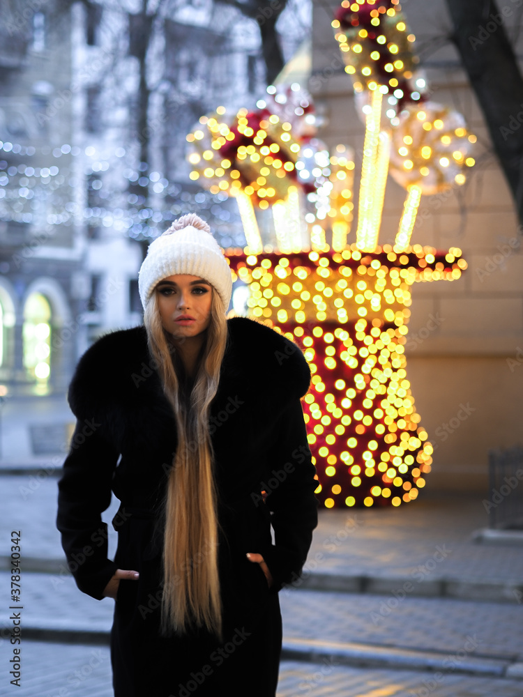 Blonde girl posing at the winter city in a black fur coat and a white hat