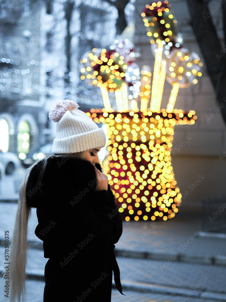 Blonde girl posing at the winter city in a black fur coat and a white hat