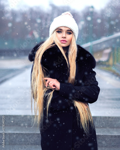 Obraz na płótnie Blonde girl posing at the winter city in a black fur coat and a white hat w salonie