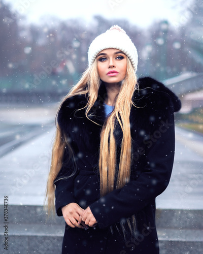 Obraz na płótnie Blonde girl posing at the winter city in a black fur coat and a white hat w salonie