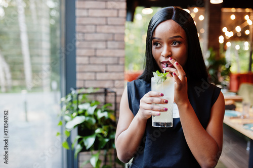 Fashionable feminist african american woman wear in black t-shirt and shorts, posed at restaurant with lemonade glass.