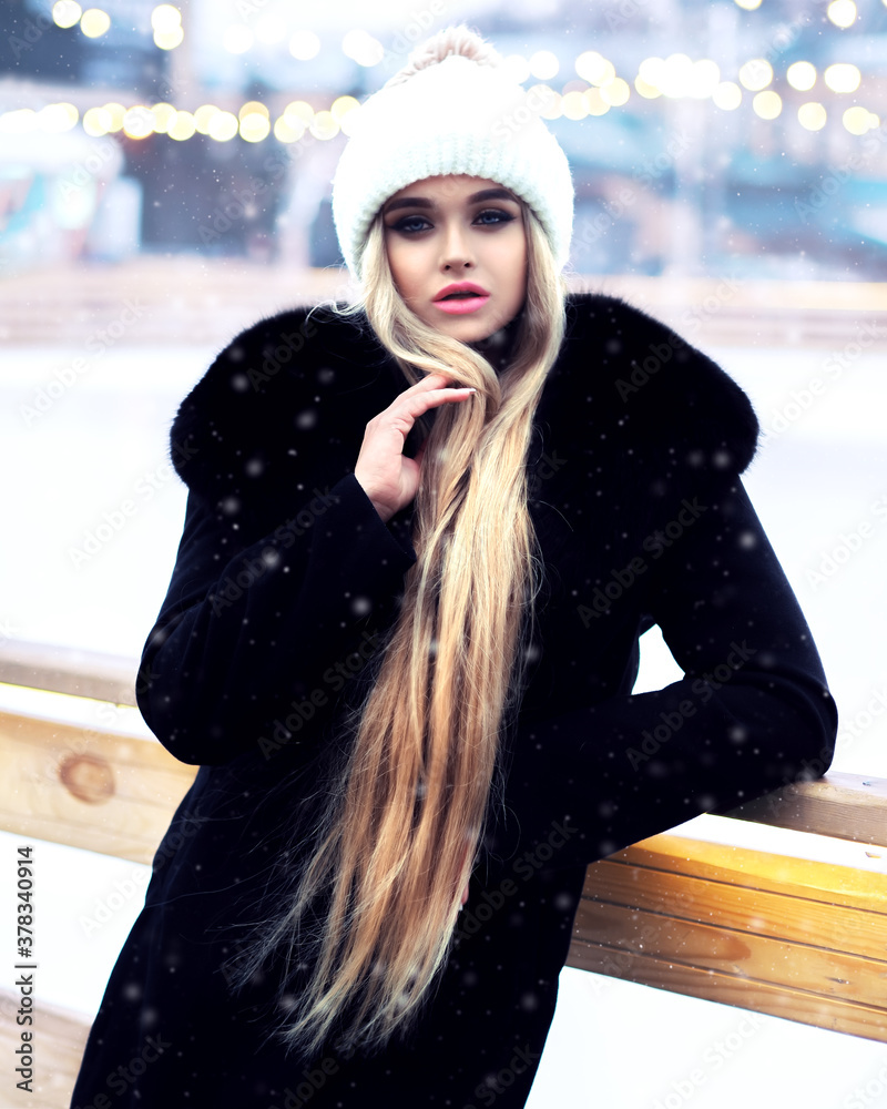 Blonde girl posing at the winter city in a black fur coat and a white hat