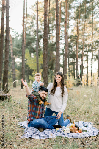 Happy family, handsome father, attractive mother and joyful little child toddler son, having fun together in autumn forest, sitting on blanket with fruits. Child is sitting on the shoulders of father