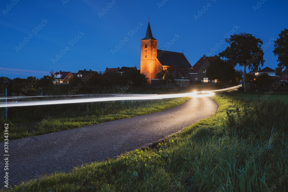 Obraz premium Car lighting streaks of light drive past a small village church in a Dutch landscape