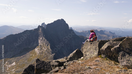 Obraz na płótnie The girl traveler sits on a high rock and admires the view of the mountains. Hiking in Ergaki. Backpack, windbreaker, boots w salonie
