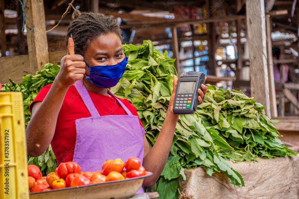 african woman in a local market wearing a face mask and holding a pos machine gives a thumbs up