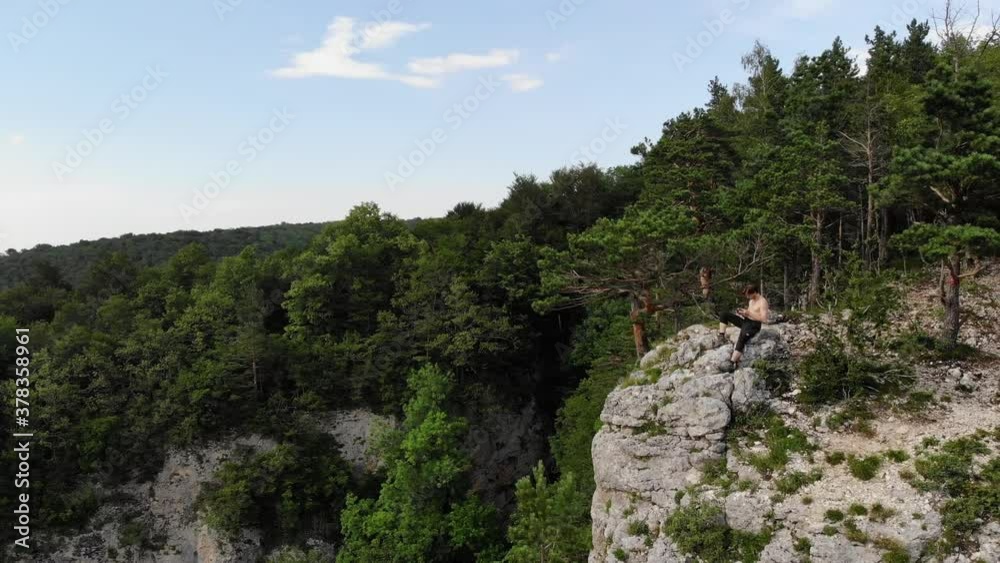 An attractive long-haired, athletic man sits on the edge of a cliff holding a drone control panel. Drone flights in nature reserves and drone recreation An attractive long-haired, athletic man sits on the edge of a cliff holding a drone control panel. Drone flights in nature reserves and drone recreation