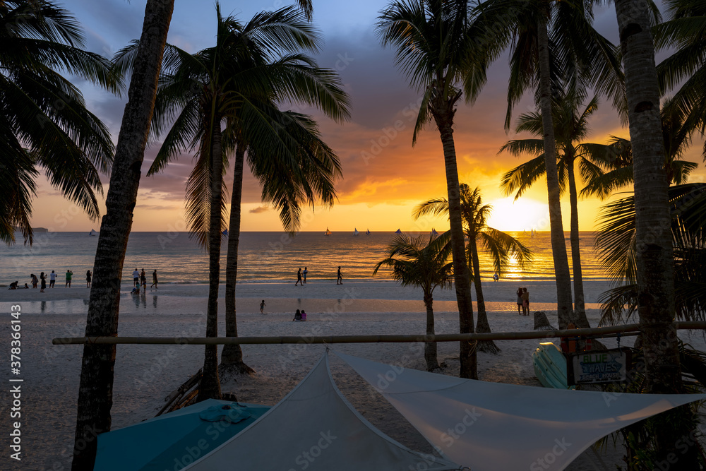 (Selective focus) Stunning view of a beautiful sunset with palm trees and the silhouette of some people relaxing on the beach. Boracay, Philippines.