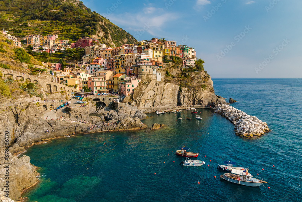 Fantastic panoramic view of the Manarola marina with anchored boats and the old colourful houses on the rocky cliff with vineyards in the background on a sunny day at the coastal area of Cinque Terre.