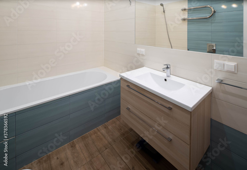 Contemporary interior of modern bathroom. White sink with wooden drawers. Huge mirror. Hardwood floor.