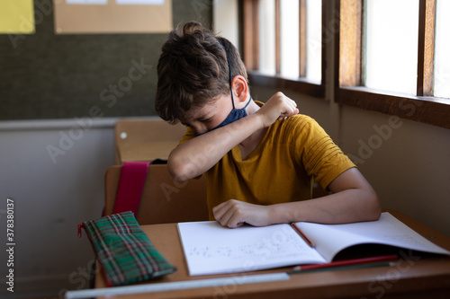 Boy wearing face mask sneezing while sitting on his desk at school 