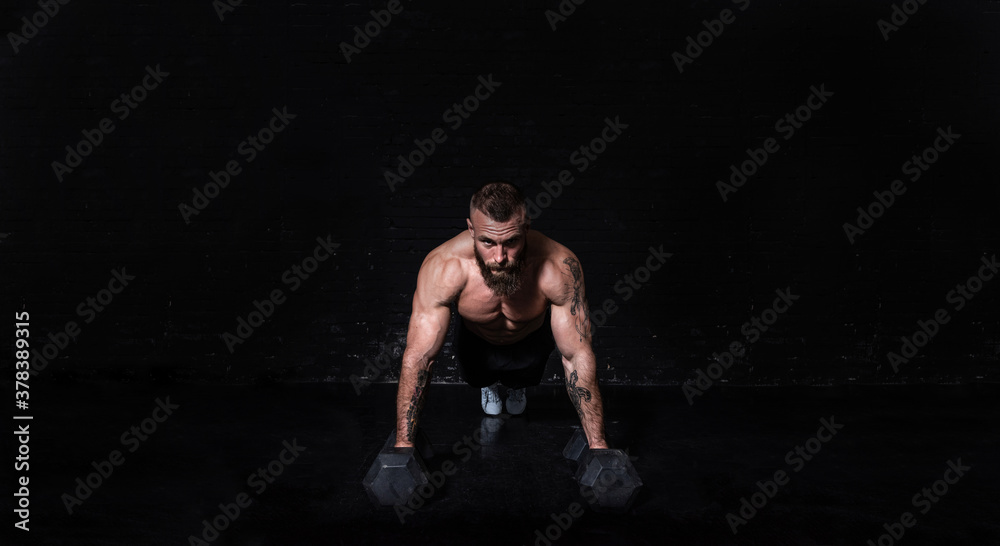 Naklejka premium Young sweaty strong fit muscular man with big muscles doing push ups on the dumbbells in the gym as hardcore cross workout training with free copy space