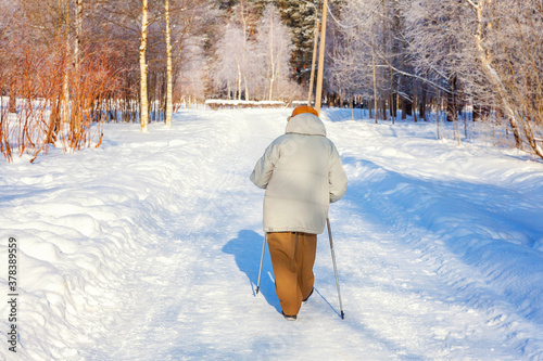 Adult woman in winter sportswear with sticks for Nordic walking on winter landscape city park background, back view. Mature woman doing exercise outdoors. Winter soprt healthy lifestyle concept photo