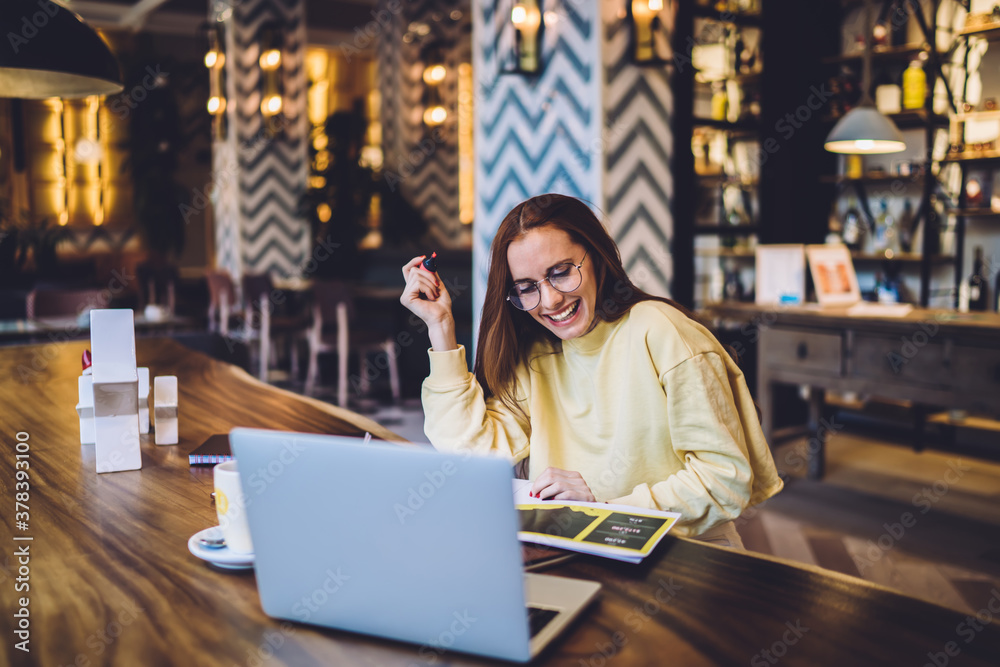 Fototapeta premium Happy Caucasian female student working on university project sitting in public cafeteria for using wifi internet and research useful information rejoicing from success,cheerful hipster girl in glasses