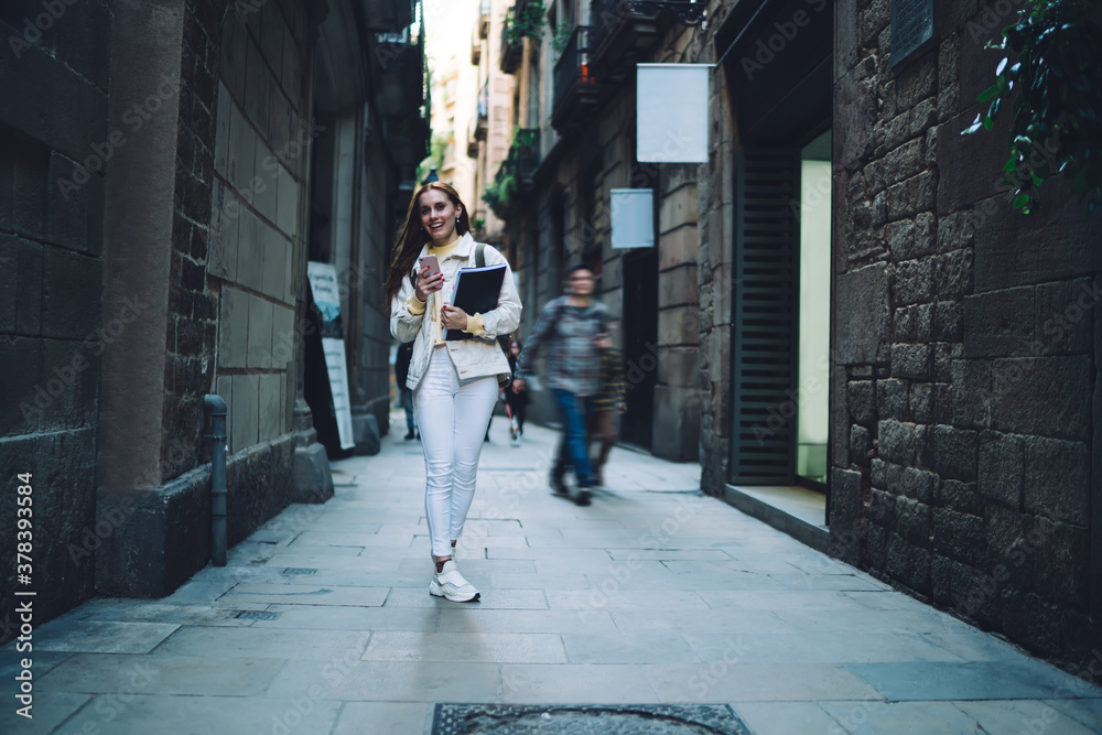Cheerful modern female student using smartphone while walking along street