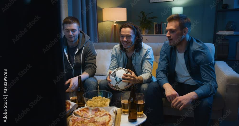 Three Caucasian cheerful best friends eating pizza and drinking beer. Guys sitting on couch at home in evening in front of TV. Football fans watching game on sport channel. Three Caucasian cheerful best friends eating pizza and drinking beer. Guys sitting on couch at home in evening in front of TV. Football fans watching game on sport channel.