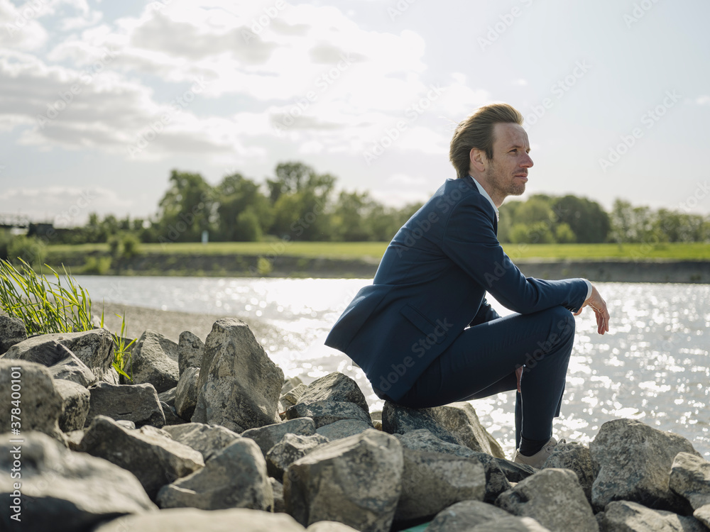 Thoughtful male entrepreneur sitting on rock at riverbank against sky Thoughtful male entrepreneur sitting on rock at riverbank against sky