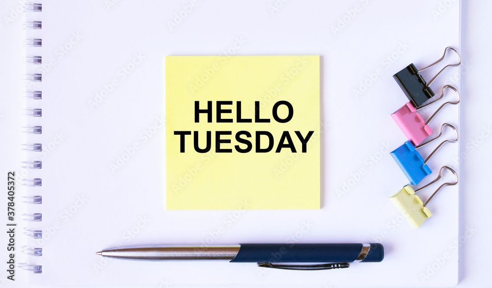 Yellow sticker with text Hello Tuesday lying on a white Notepad with a pen and paper clips Yellow sticker with text Hello Tuesday lying on a white Notepad with a pen and paper clips
