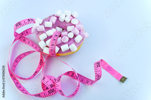 Sweet donut with icing and marshmallow on a white background with pink centimeter tape. Junk food. Diet, weight loss and health concept, measuring tape. Unhealthy eating. A bomb of calories. 